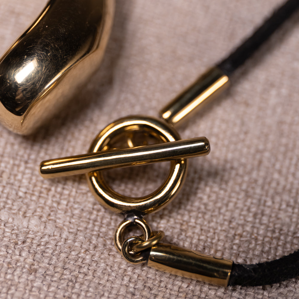 Close-up of a gold-tone toggle ring clasp with a black cord, resting on a textured beige fabric surface.