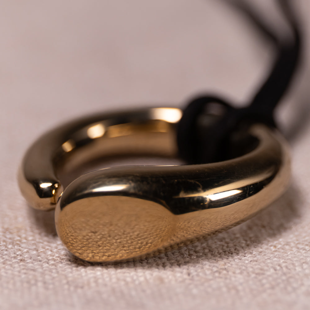 A close-up of a polished gold-toned ring with a textured front face and a black cord attached, resting on a light beige fabric surface.