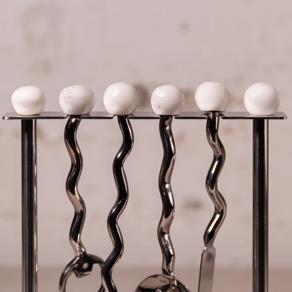 A close-up of a metal utensil holder with five white spherical knobs on a black top bar, holding silver kitchen utensils against a soft beige background.