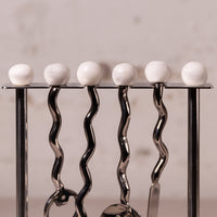 A close-up of a metal utensil holder with five white spherical knobs on a black top bar, holding silver kitchen utensils against a soft beige background.