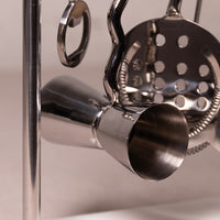 Close-up of a polished stainless steel cocktail jigger and matching metal bar tools hanging on a stand.