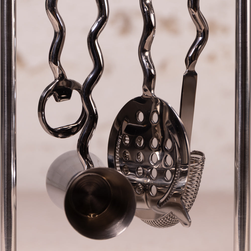 A close-up of a stainless steel cocktail tool set hanging from wavy metal hooks, including a strainer, muddler, and bottle opener, against a softly blurred light background.
