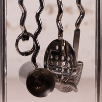 A close-up of a stainless steel cocktail tool set hanging from wavy metal hooks, including a strainer, muddler, and bottle opener, against a softly blurred light background.