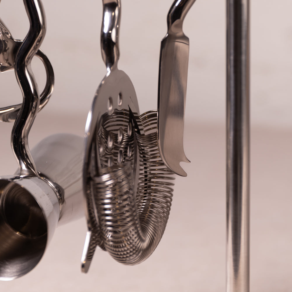 Close-up of stainless steel cocktail bar tools hanging from a metal stand, including a strainer, spoon, and other bar accessories.
