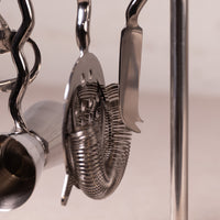 Close-up of stainless steel cocktail bar tools hanging from a metal stand, including a strainer, spoon, and other bar accessories.
