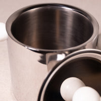 Close-up of a brushed stainless steel cylindrical container with a matching metal lid resting beside it on a light neutral surface.
