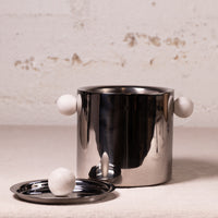 A polished stainless steel cylindrical container with two white spherical handles and a matching small metal tray, photographed against a light textured wall.