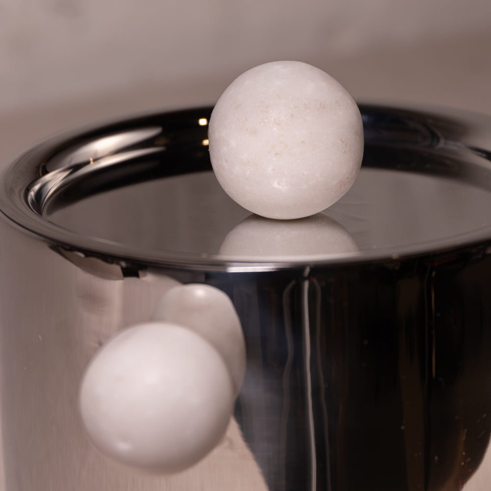 A close-up of a polished metallic pot or container with two smooth white spherical objects resting on and near its reflective surface.
