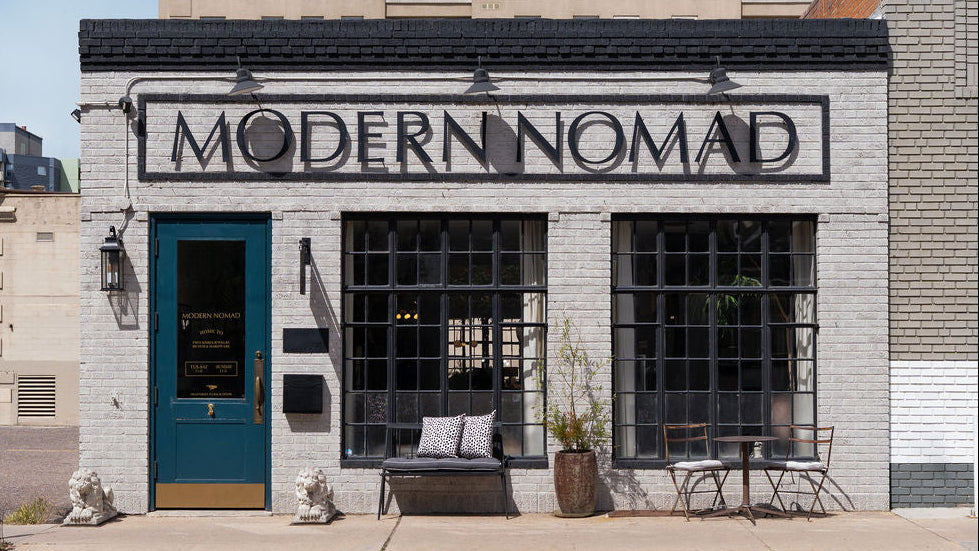 The exterior of a brick building featuring a large black sign that reads 'MODERN NOMAD' above a teal door and two large black-framed windows. A bench with pillows, a potted plant, and small outdoor seating sit on the sidewalk in front.