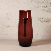 A tall, dark amber-brown translucent glass pitcher with a handle, standing on a light beige surface against a textured off-white wall.