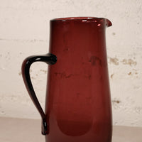 A translucent dark red glass pitcher with a curved black handle, shown against a textured off-white brick wall background.