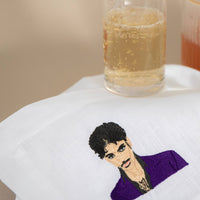 A white napkin featuring an embroidered portrait of Prince with a purple jacket, styled on a beige surface with a glass of bubbly drink in the background.