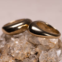 Two polished gold rings with a unique rounded, organic shape resting on a bed of clear quartz crystals against a neutral beige background.