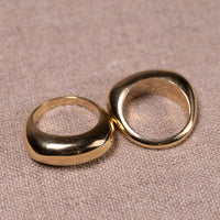 Two thick, chunky gold rings with organic, rounded shapes resting on a textured beige fabric background.