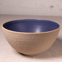 A handmade ceramic bowl with a textured beige exterior and a smooth, glossy blue interior, set against a neutral light gray background.