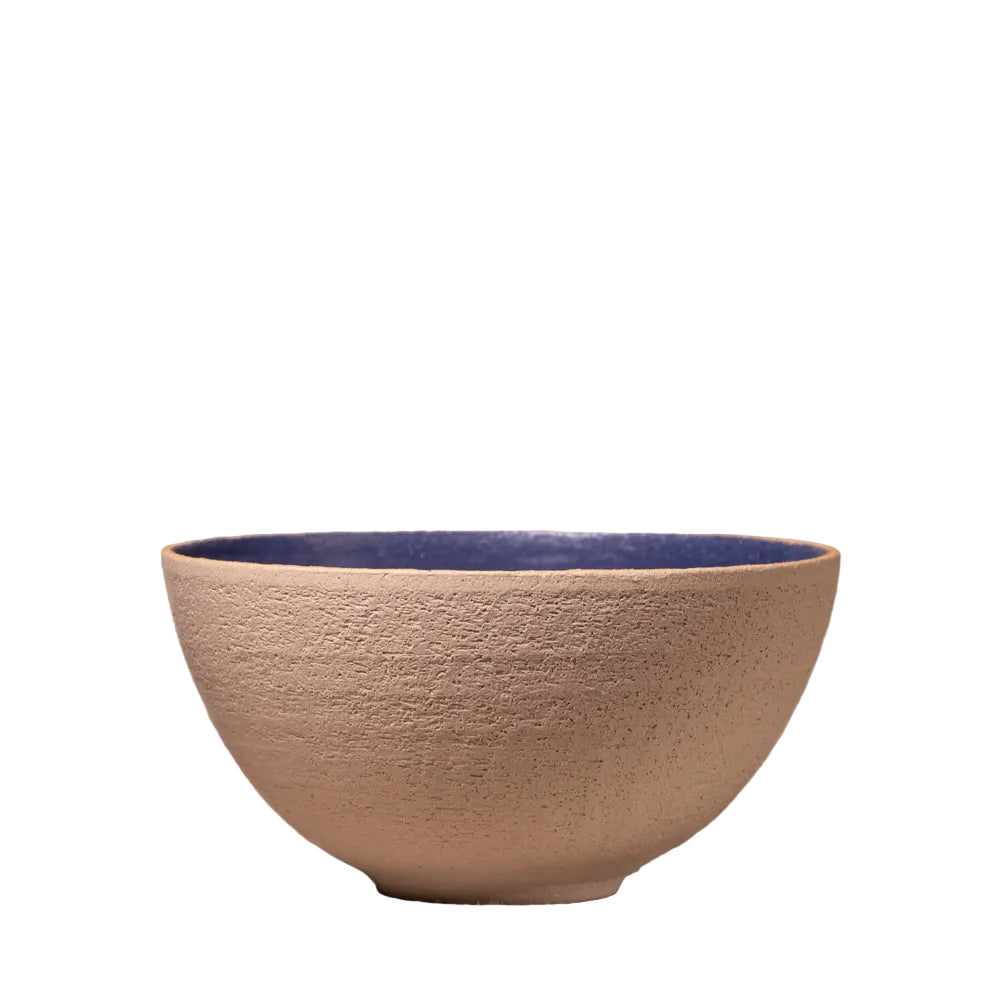 A handmade ceramic bowl with a textured beige exterior and a glossy dark blue interior, set against a rustic, light-colored brick wall background.