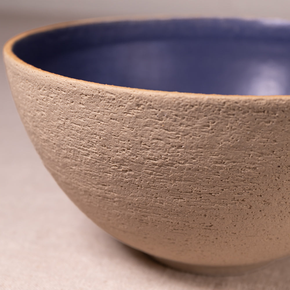 Close-up of a ceramic bowl with a textured beige exterior and a smooth dark blue interior, shown on a neutral light gray surface.