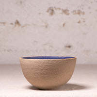 A handmade ceramic bowl with a textured beige exterior and a contrasting blue glazed interior, set against a rustic, light-colored plaster wall.