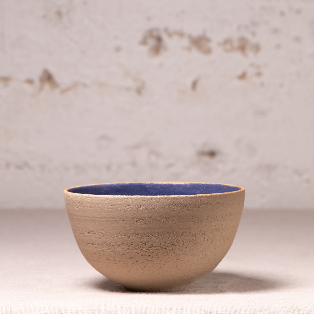 A handmade ceramic bowl with a textured beige exterior and a contrasting dark blue glazed interior, set against a neutral, light-colored rustic background.