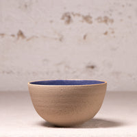A handmade ceramic bowl with a textured beige exterior and a contrasting dark blue glazed interior, set against a neutral, light-colored rustic background.