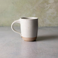 A handmade ceramic mug with a matte off-white body, a natural tan unglazed base, and a curved handle, set against a textured light gray and green background.